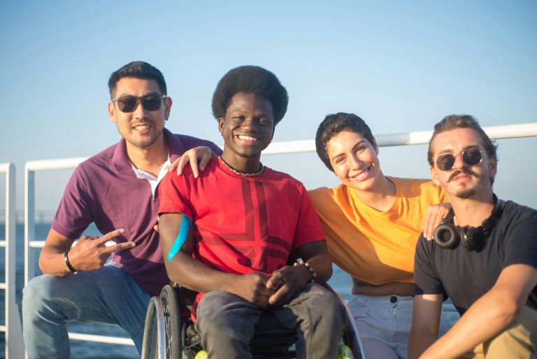 A black man in a red t-shirt is in a wheel chair and is surrounded by three of his friends, two man and one woman, who are all of different races and ethnicities. They are outside near the water enjoying a sunny day.