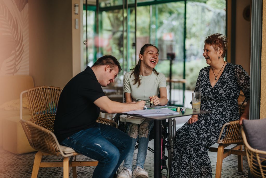Image of three people, a young woman, young man and middle aged-woman, siting around a table laughing. The young man and woman are drawing.