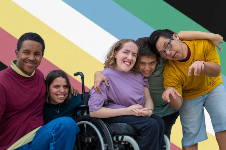 A group of five young adults with diverse disabilities smiling in front of a disability pride flag